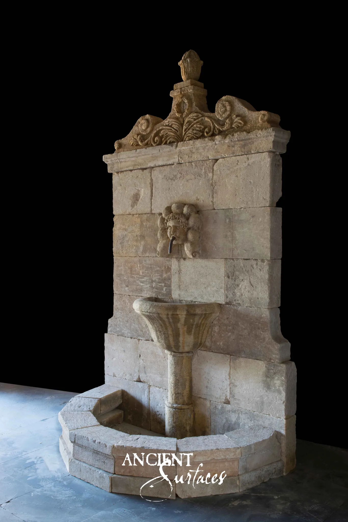 Tall reclaimed antique limestone wall fountain from Ancient Surfaces with a carved acanthus pediment, mask spout and stepped semicircular basin