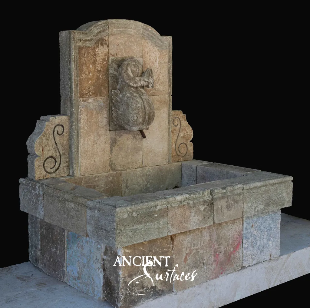 Reclaimed antique limestone wall fountain from Ancient Surfaces carved with a coiled dolphin above a deep square block basin